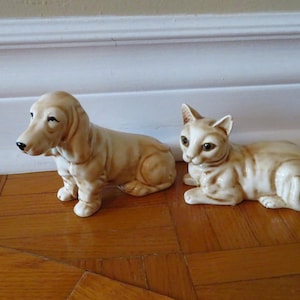 Peut inclure: Deux figurines d'animaux en céramique : un chien teckel et un chat. Le chien est beige clair avec un long corps et des oreilles tombantes. Le chat est également beige clair avec des rayures plus foncées et est couché.