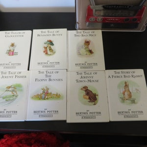 May include: A collection of eight vintage children's books by Beatrix Potter. The books feature illustrations of various characters, including Peter Rabbit, Jeremy Fisher, and Benjamin Bunny. The books are arranged in two rows of four on a black surface.