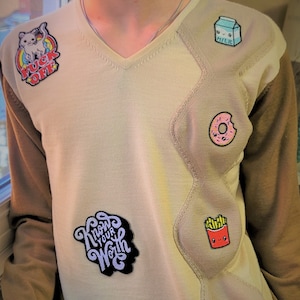 May include: A beige sweater with a v-neck and brown sleeves. The sweater has several embroidered patches, including a cartoon pig with a rainbow, a milk carton, a doughnut, chips, a hamburger, and a patch that says "Know Your Worth".