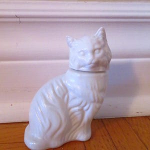 Pode incluir: Estatueta de gato em cerâmica branca com cabeça removível, possivelmente um recipiente decorativo. O gato está sentado com textura de pelo detalhada. A estatueta está colocada num chão de madeira.