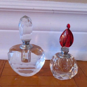 Puede incluir: Dos frascos de perfume de vidrio transparente con tapones facetados. El frasco más grande tiene un tapón transparente, mientras que el frasco más pequeño tiene un tapón de vidrio rojo.
