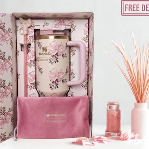 May include: A cream-colored Stanley tumbler with a pink floral and bow pattern, featuring a matching pink handle, a clear lid, and a pink straw with a bow topper. It is presented in a matching patterned box with a pink velvet pouch and a pink logo.
