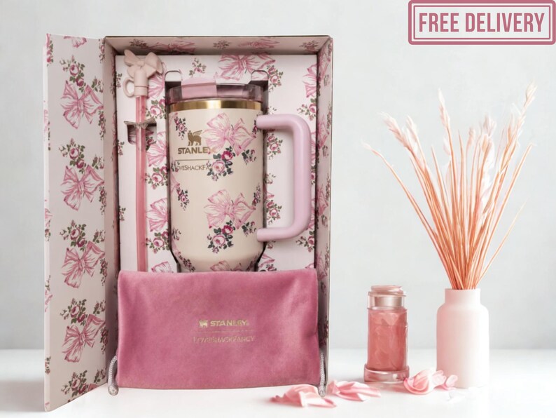 May include: A cream-colored Stanley tumbler with a pink floral and bow pattern, featuring a matching pink handle, a clear lid, and a pink straw with a bow topper. It is presented in a matching patterned box with a pink velvet pouch and a pink logo.