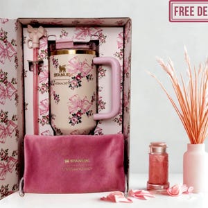 May include: A cream-colored Stanley tumbler with a pink bow and floral pattern, featuring a matching pink straw with a bow topper, housed in a decorative box. A dark pink velvet pouch with the Stanley logo sits in front of the tumbler inside the box.