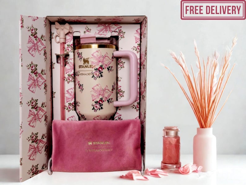 May include: A cream-colored Stanley tumbler with a pink bow and floral pattern, featuring a matching pink straw with a bow topper, housed in a decorative box. A dark pink velvet pouch with the Stanley logo sits in front of the tumbler inside the box.