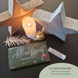 May include: A festive arrangement featuring a lit candle, gold ornament, and a chocolate bar with the text "Adventszeit". Two star-shaped decorations and a paper cutout add to the holiday theme. The image also includes a small scroll with text.