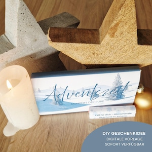Peut inclure: Une bougie blanche allumée, une boîte avec le mot "Adventszeit" et un petit rouleau avec du texte, et un ornement doré sont sur une surface en bois. Une grande étoile en bois et une étoile en béton sont en arrière-plan. La boîte a une scène d'hiver.