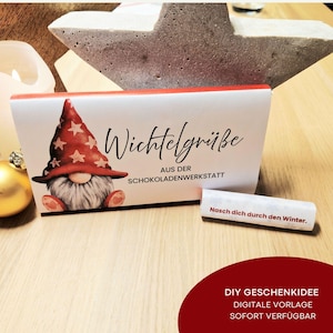 Może przedstawiać: Białe pudełko z czerwonymi obramowaniami z ilustracją gnoma w czerwonym kapeluszu z białymi gwiazdami. Tekst na pudełku brzmi "Wichtelgrüße" i "AUS DER SCHOKOLADENWERKSTATT". Mały zwój z tekstem "Nasch dich durch den Winter."