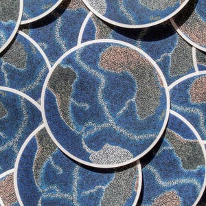 May include: A collection of round stickers with a blue, brown, and gray abstract design. Each sticker has a white border. The design features a dotted pattern, resembling a map or geographical representation.