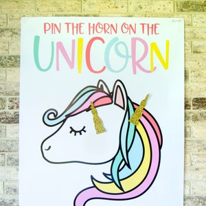 Puede incluir: Un póster blanco con el texto "Pin the horn on the unicorn" en letras de colores. Un unicornio de dibujos animados con una melena y una cola arcoíris está en el póster. El unicornio tiene los ojos cerrados y un cuerno dorado.