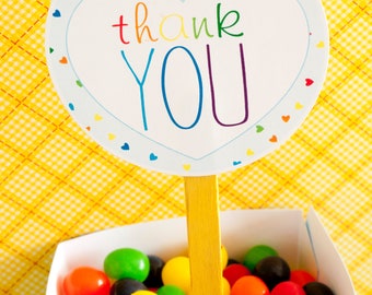 May include: A white sign with the words "thank YOU" in rainbow colors, held by a yellow stick, sits above a box filled with colorful jelly beans. The sign has a heart-shaped border with small heart accents.