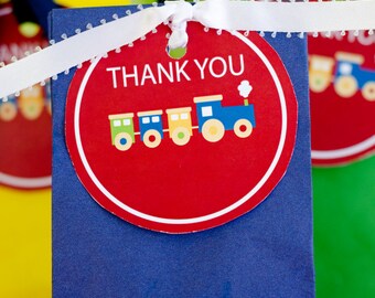 May include: A navy blue gift bag with a red tag that says "THANK YOU" and features a colorful train illustration. The tag is tied with a white ribbon. Other gift bags are visible in the background.