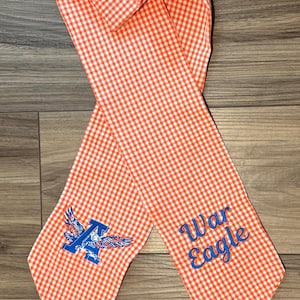 May include: An orange and white gingham fabric scarf featuring an embroidered blue letter A with eagle wings on one end and the blue embroidered text War Eagle on the other end. The scarf has pointed ends and a long, narrow, rectangular shape.