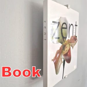 Puede incluir: Un libro blanco con la palabra "zent" en el lomo y "zent" en la portada, con una ilustración floral. El libro está montado en una pared con una exhibición de libros flotante. Las palabras "Floating Book Display" son rojas.