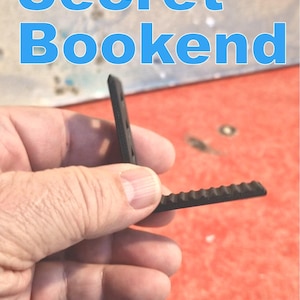 May include: A black, plastic bookend held in a hand. The bookend has a ridged edge and a vertical support. The text "Secret Bookend" is in blue letters at the top of the image.