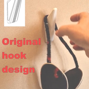 May include: A white wall hook with a black and red strap holding a pair of black and white ear muffs with a red cherry pattern. The text "Original hook design" is in red.