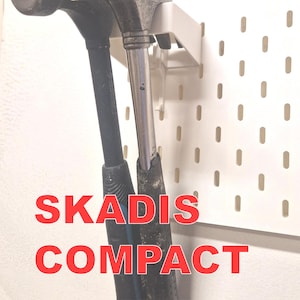 Peut inclure: Deux marteaux, un noir et un argenté, sont montés sur un panneau perforé blanc. Le texte "SKADIS COMPACT HAMMERS" est superposé en rouge. Les marteaux sont maintenus par un support blanc.