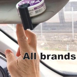 Könnte beinhalten: Ein schwarzer Clip hält einen weißen und lila Behälter mit der Aufschrift "VELO" und "GROOVY GRAPE" an der Sonnenblende eines Autos fest. Der Text "All brands" wird in schwarzen Buchstaben angezeigt.