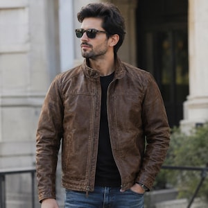 May include: A brown leather jacket with a stand-up collar, a full-length front zipper, and long sleeves. The jacket features a distressed finish, horizontal seam detailing across the chest, and two side pockets. It is worn over a black shirt.