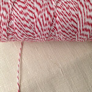May include: A spool of red and white baker's twine. The twine is tightly wound, with a single strand extending from the spool. The background is a beige fabric.