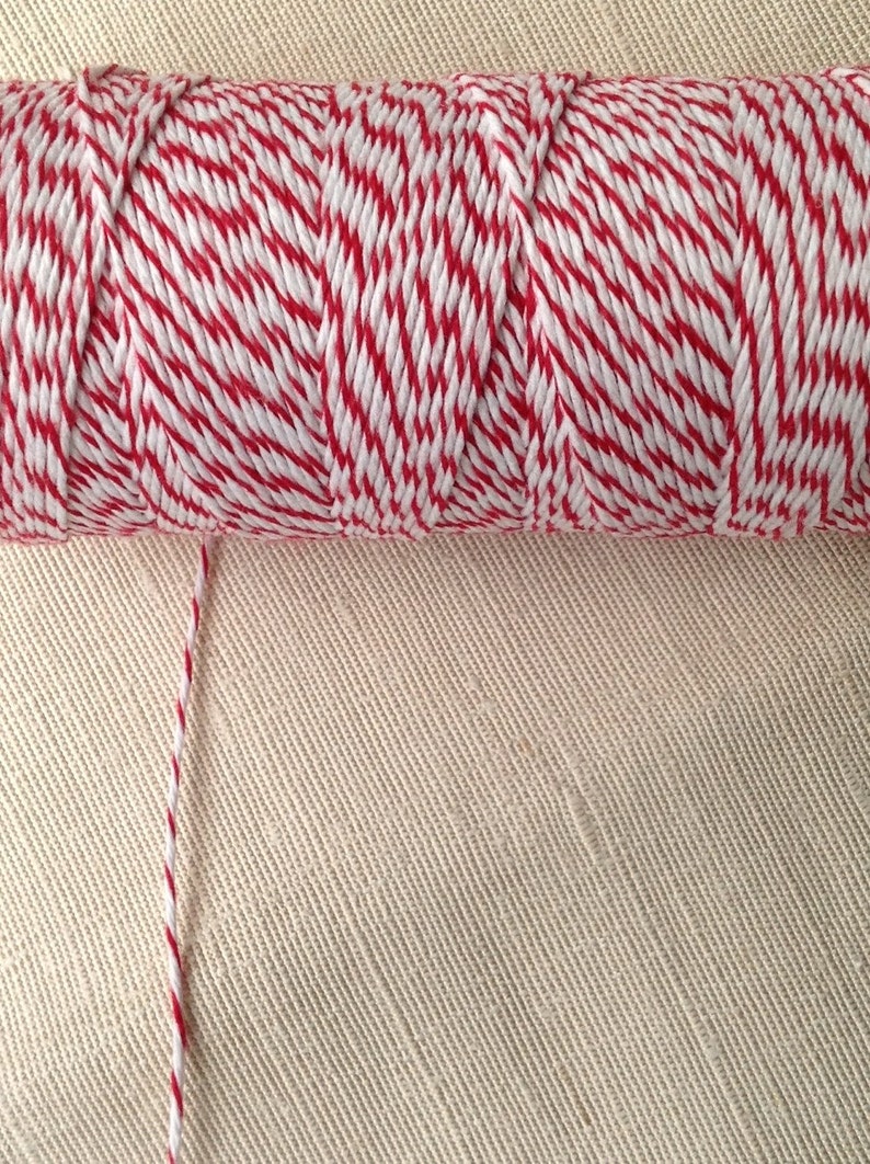 May include: A spool of red and white baker's twine. The twine is tightly wound, with a single strand extending from the spool. The background is a beige fabric.