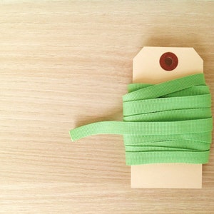 May include: A roll of light green ribbon is attached to a beige tag with a brown circular accent. The ribbon is neatly coiled, with a small portion extending outwards. The background is a light wood grain.