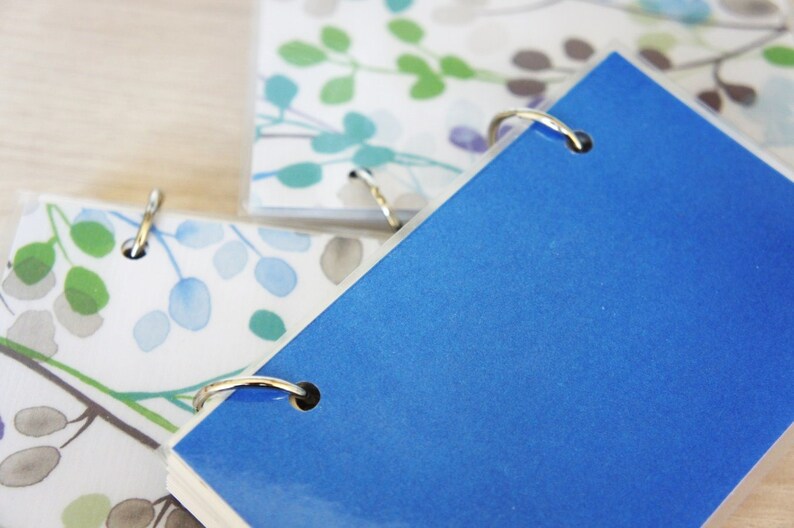 May include: Close-up of several small notebooks with metal ring bindings. The top notebook is a solid blue, while others display a watercolor floral pattern in shades of green, blue, and gray. The notebooks are on a light wooden surface.