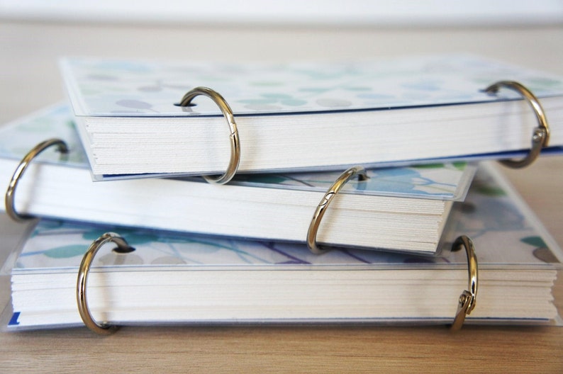 May include: Several small notebooks with white pages and floral patterned covers are stacked. Each notebook is bound with a gold-colored metal ring. The covers feature a blue and green floral design. The notebooks are on a light-colored wooden surface.