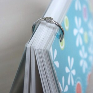 May include: A notebook with a light blue cover, white floral pattern, and a silver binder ring. The pages are white and the notebook is partially open, revealing the edge of the pages.