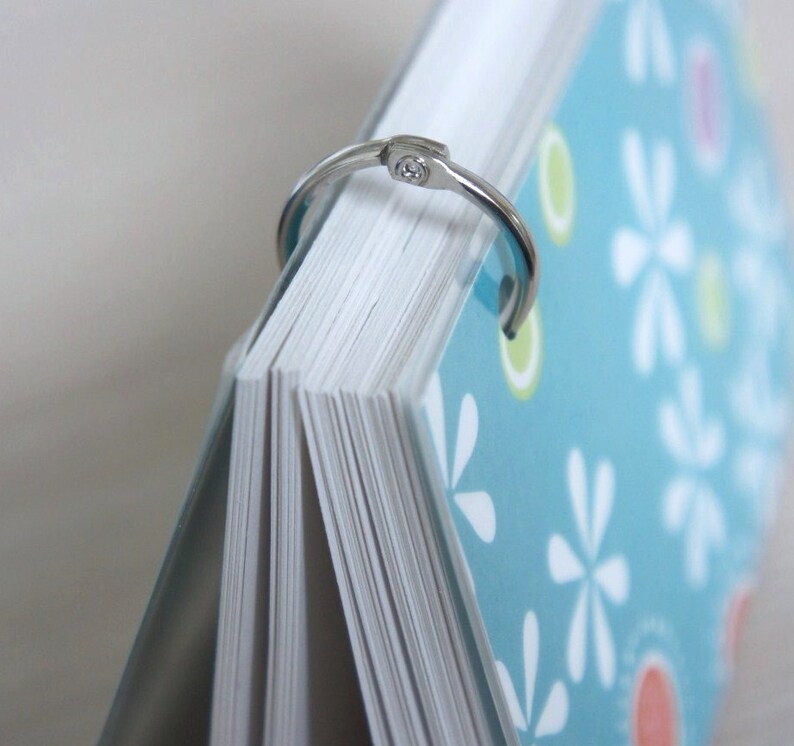 May include: A notebook with a light blue cover, white floral pattern, and a silver binder ring. The pages are white and the notebook is partially open, revealing the edge of the pages.
