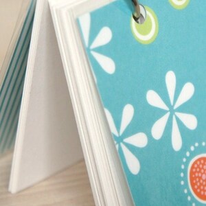 May include: A small notebook with a teal cover featuring white flower and dot patterns. The pages are held together by a silver ring. The notebook's pages are white and the cover has a clear plastic overlay.