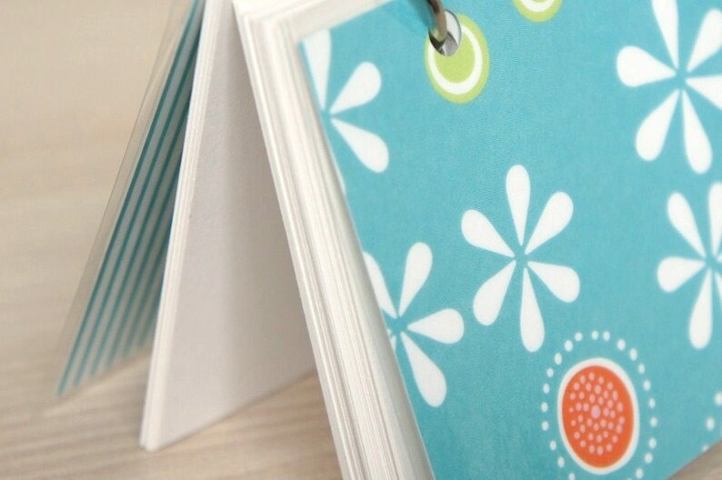 May include: A small notebook with a teal cover featuring white flower and dot patterns. The pages are held together by a silver ring. The notebook's pages are white and the cover has a clear plastic overlay.