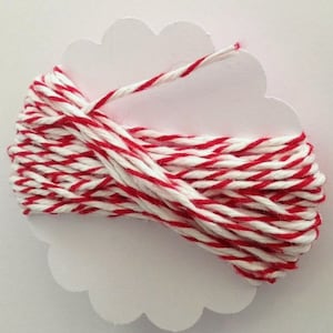 May include: A spool of red and white striped twine is wrapped around a white, cloud-shaped card. The twine is tightly wound, with the red stripes evenly spaced among the white. The card provides a clean, neutral backdrop for the colorful twine.