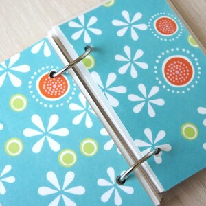May include: Open notebook with a teal cover featuring a white floral pattern, orange and white circular designs, and green circles. The notebook is held together by silver metal rings.