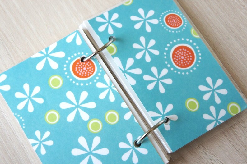 May include: Open notebook with a teal cover featuring a white floral pattern, orange and white circular designs, and green circles. The notebook is held together by silver metal rings.