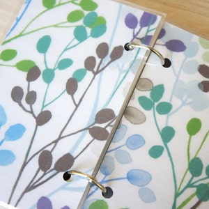 May include: Two ring-bound notebooks with a white background and a repeating pattern of stylized leaves and branches in shades of green, blue, and purple. The metal rings are silver.