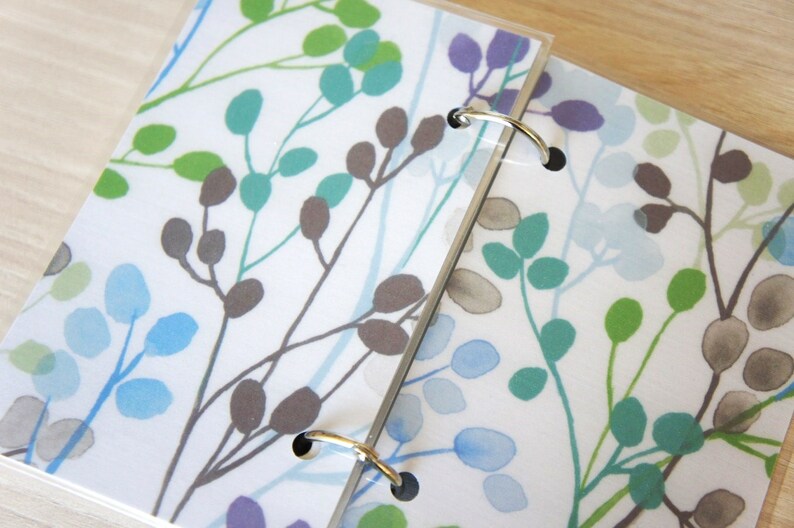 May include: Two ring-bound notebooks with a white background and a repeating pattern of stylized leaves and branches in shades of green, blue, and purple. The metal rings are silver.