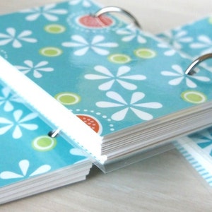 May include: A collection of small notebooks featuring a floral pattern. The covers are teal with white daisy-like flowers, green circles, and orange details. The pages are bound with silver metal rings.