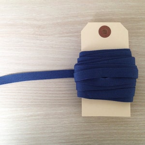 May include: A spool of navy blue grosgrain ribbon, partially unrolled, sits on a light-colored tag. The ribbon is tightly wound, with a textured surface. A small, round, brown sticker is affixed to the tag.