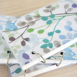 May include: A stack of small notebooks with clear plastic covers. The covers feature a watercolor floral design in shades of blue, green, and gray. The notebooks are bound with silver metal rings.