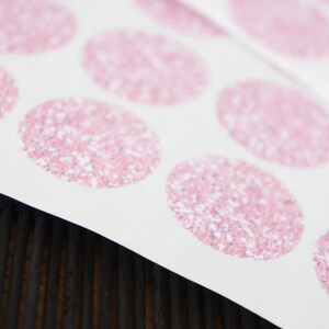 May include: Sheet of pink glitter circle stickers on white paper. The stickers are arranged in rows and columns. The background is a dark brown wooden surface.