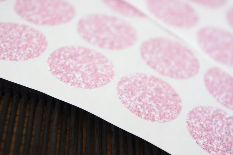 May include: Sheet of pink glitter circle stickers on white paper. The stickers are arranged in rows and columns. The background is a dark brown wooden surface.