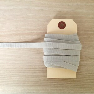 May include: A spool of light gray grosgrain ribbon, partially unwound, is displayed on a beige tag. The ribbon is neatly coiled, with a portion extending to the left. The background is a light wood grain.