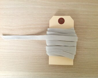 May include: A spool of light gray grosgrain ribbon, partially unwound, is displayed on a beige tag. The ribbon is neatly coiled, with a portion extending to the left. The background is a light wood grain.