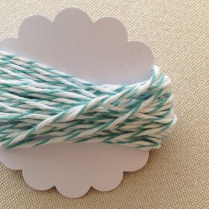 May include: A spool of teal and white striped twine sits on a white, scalloped-edge card. The twine is tightly wound, showing the alternating colors. This craft supply is ideal for various DIY projects and gift wrapping.