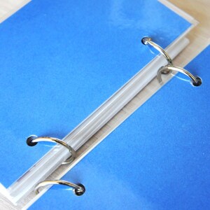 May include: Close-up of two blue binder pages with silver metal rings. The pages are a vibrant blue color and are held together by the rings. The binder is on a light-colored surface.