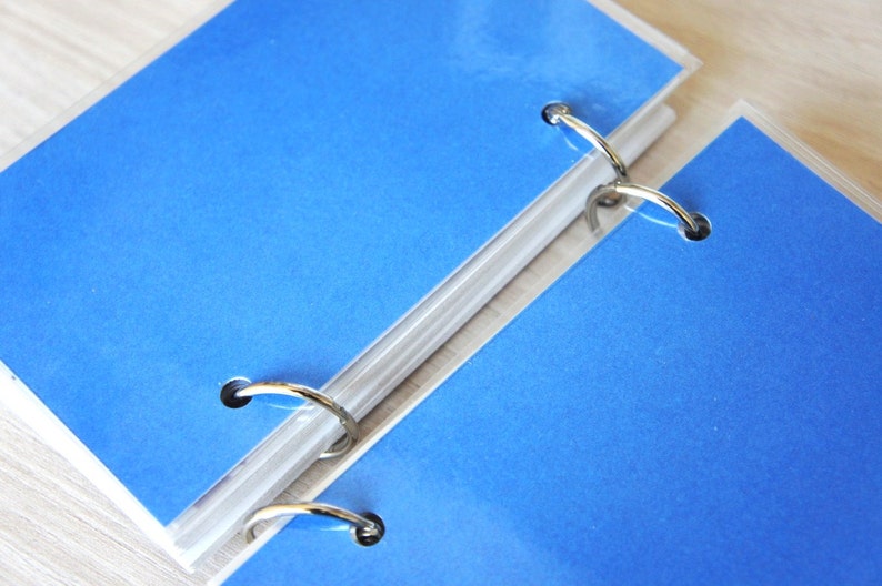 May include: Close-up of two blue binder pages with silver metal rings. The pages are a vibrant blue color and are held together by the rings. The binder is on a light-colored surface.