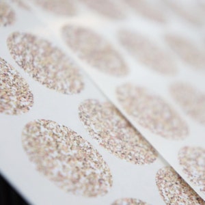 May include: Close-up of a sheet of white paper with multiple oval stickers. Each sticker has a speckled, glittery design in beige, brown, and gold. The stickers are arranged in neat rows and columns.