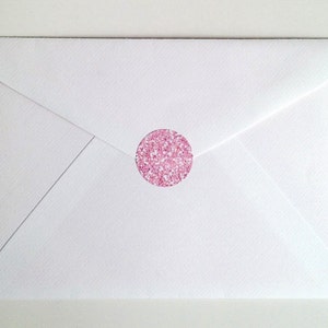 May include: A white envelope sealed with a round pink glitter sticker. The envelope is closed and has a triangular flap. The sticker is centered on the envelope and has a sparkly appearance.