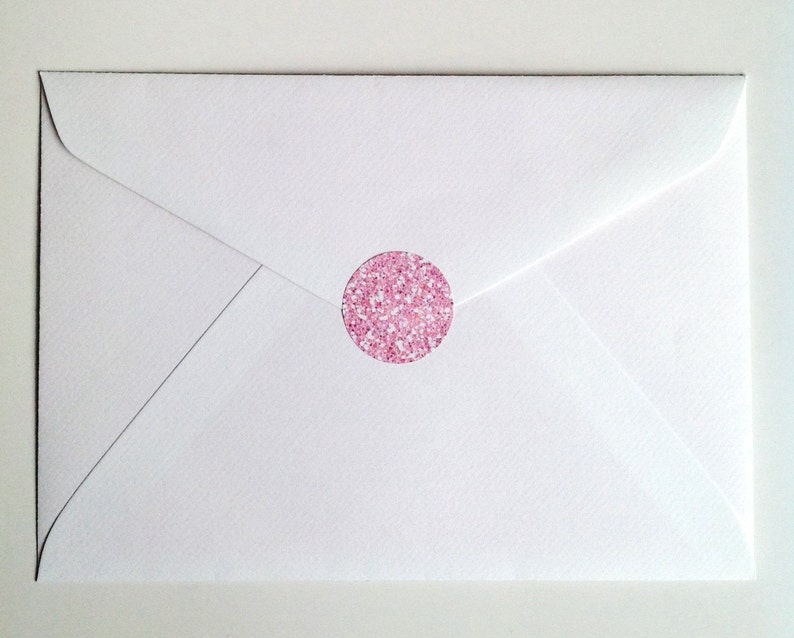 May include: A white envelope sealed with a round pink glitter sticker. The envelope is closed and has a triangular flap. The sticker is centered on the envelope and has a sparkly appearance.